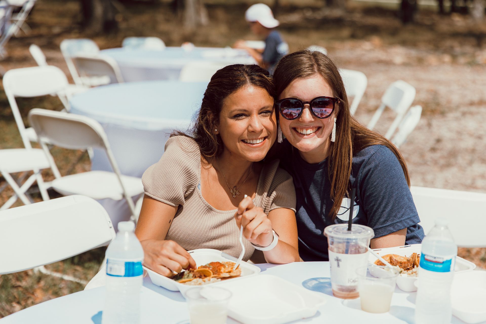Two people smile at a table at an outdoor event, eating from plates with bottled water nearby.