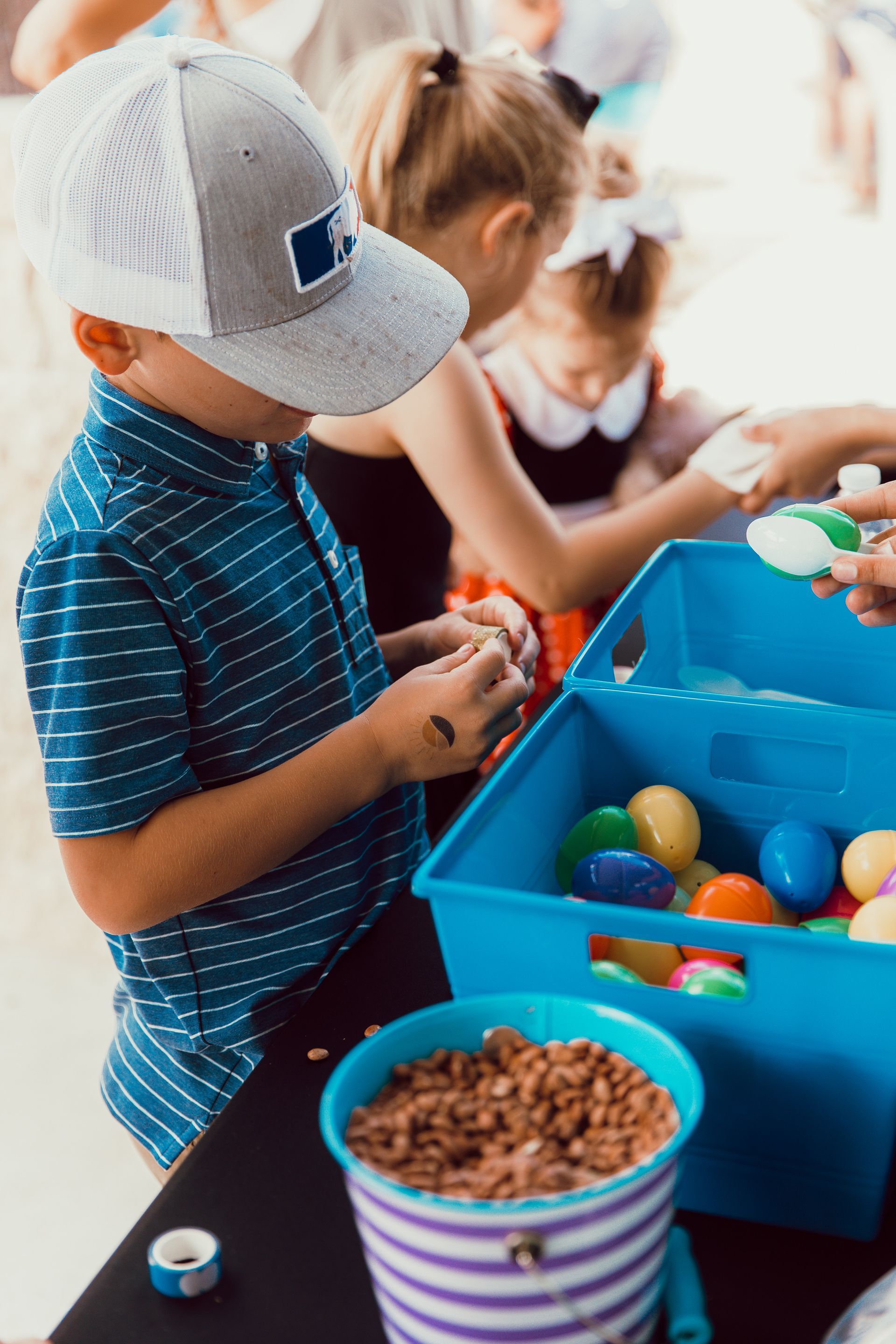 Children in a group reach for colorful plastic eggs from a blue bin during an outdoor activity.