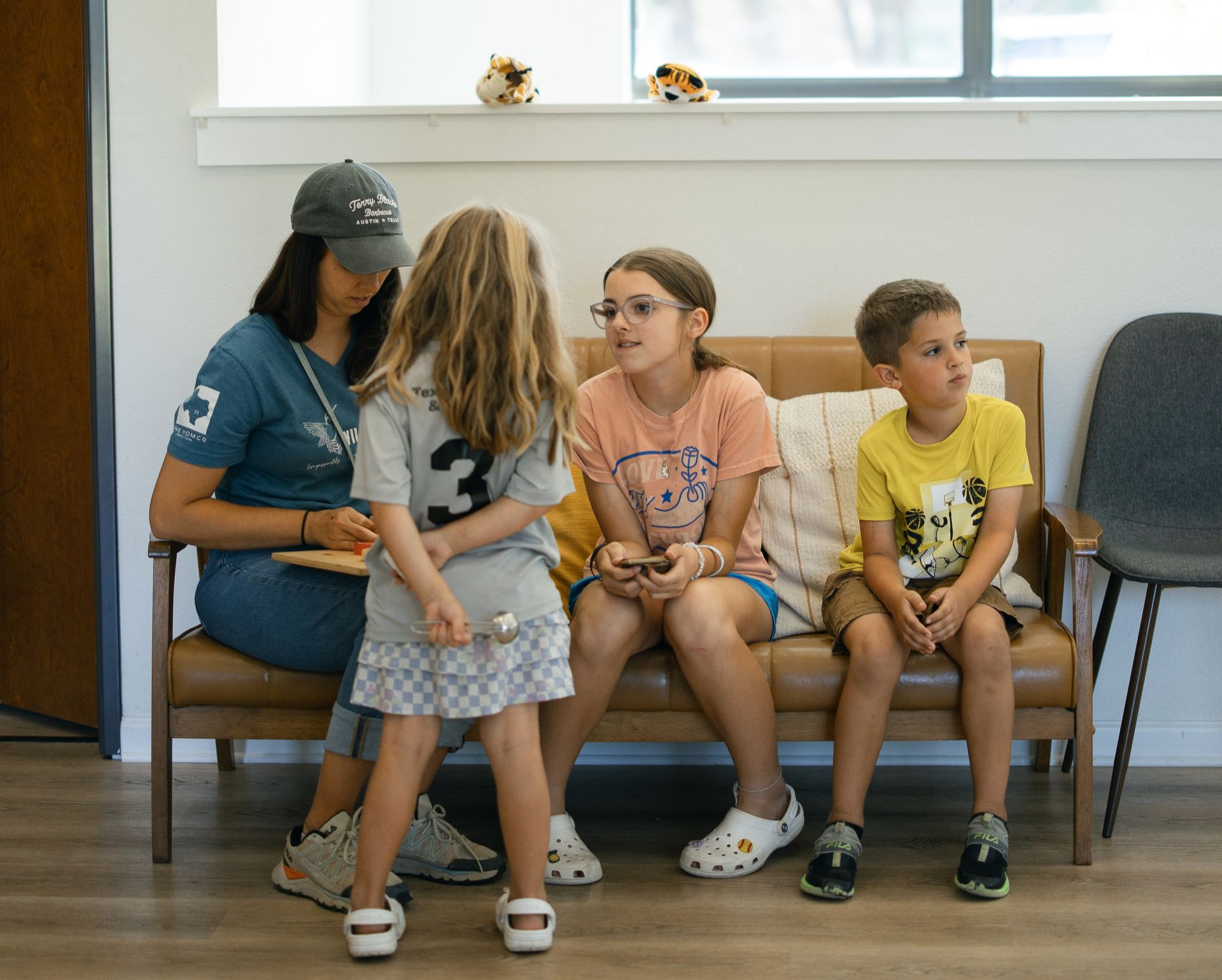 Woman and three children seated on a brown bench, talking. Brightly lit room.