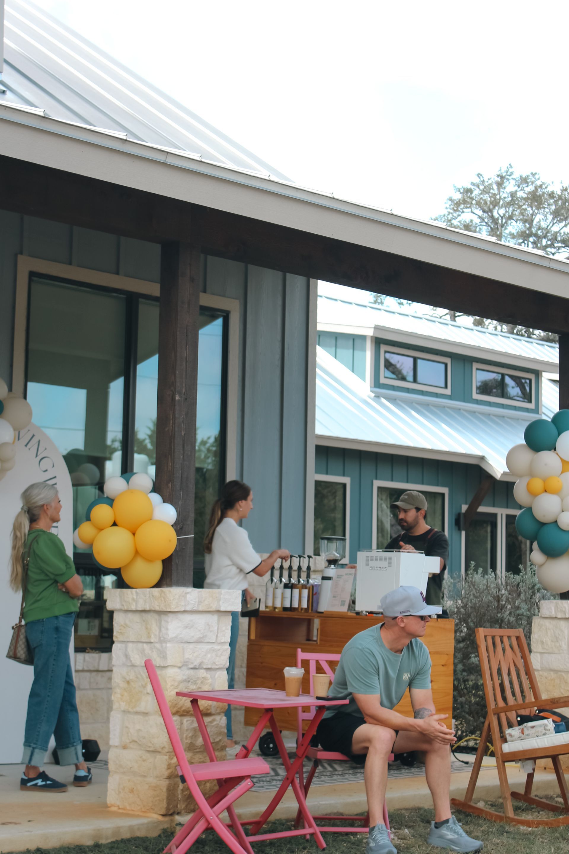 People gather at a patio outside a blue house, decorated with balloon garlands and a small serving station.