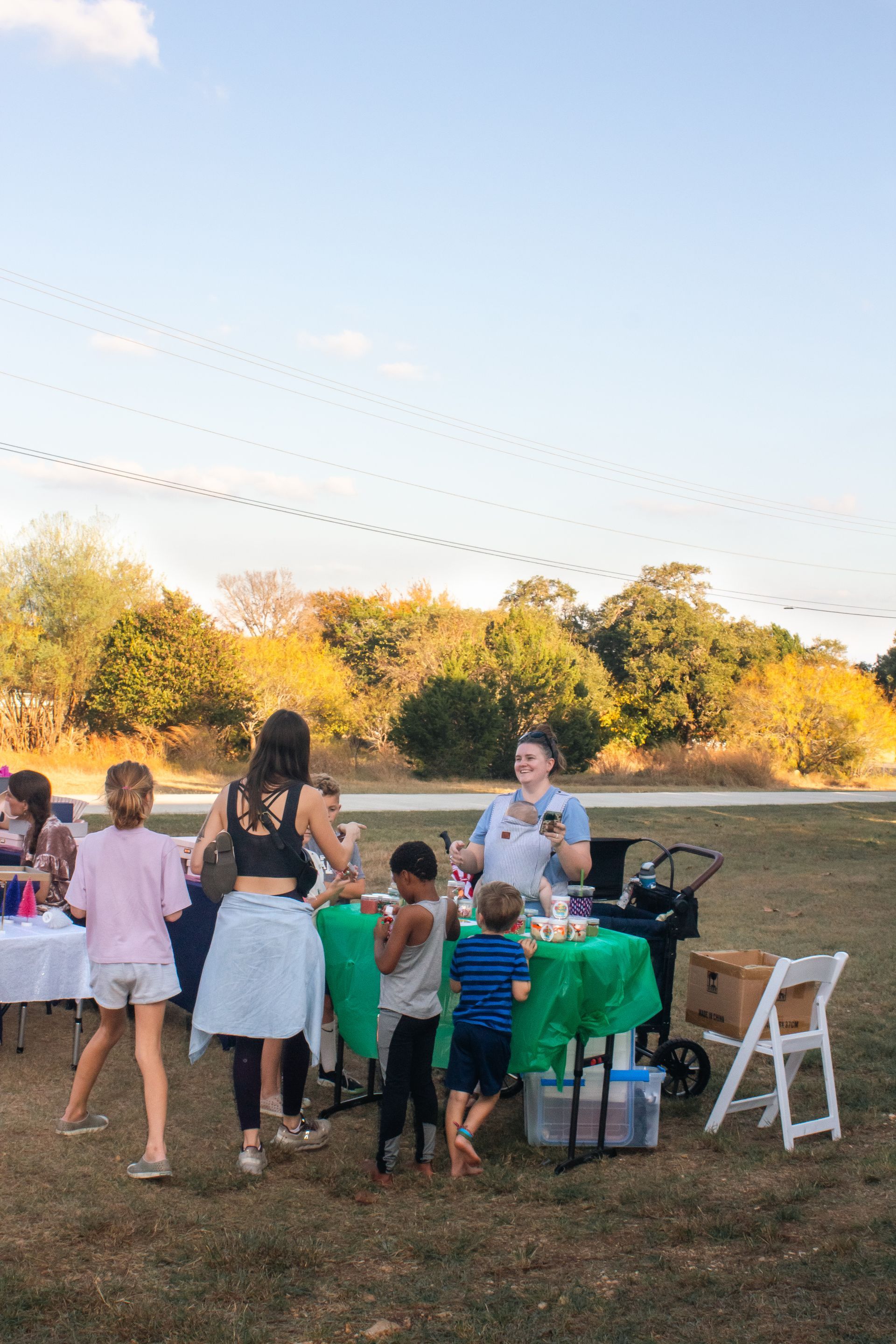 People gather at an outdoor event with tables set up on a grassy field under a sunny sky.