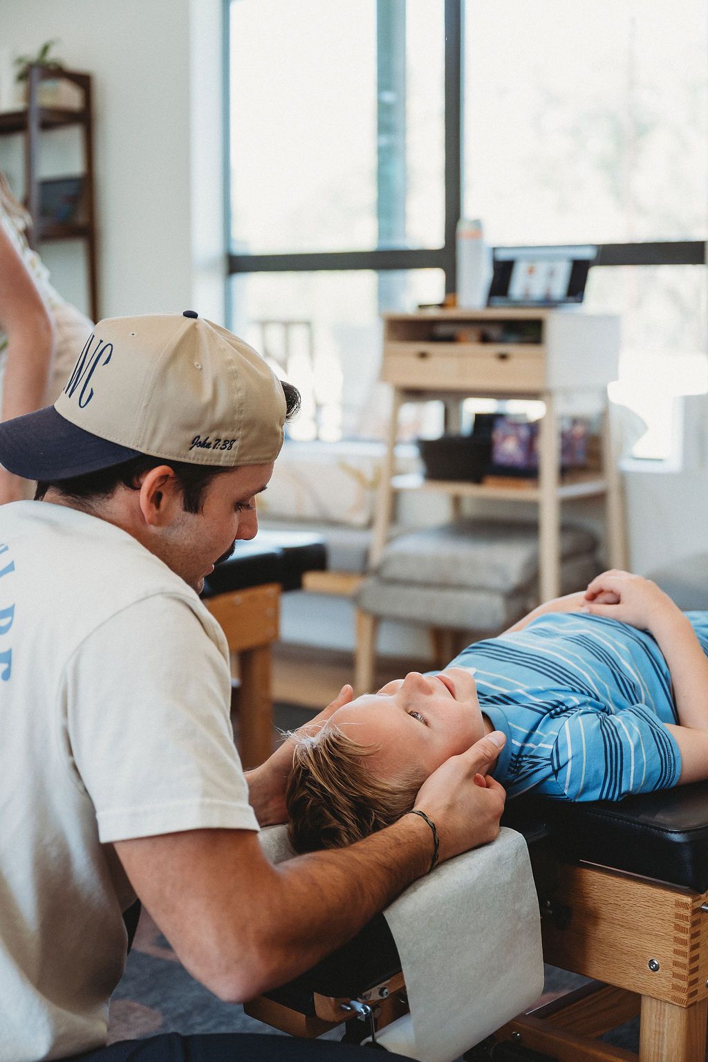 A chiropractor adjusts the head and neck of a child lying on a treatment table in a bright, modern clinic.