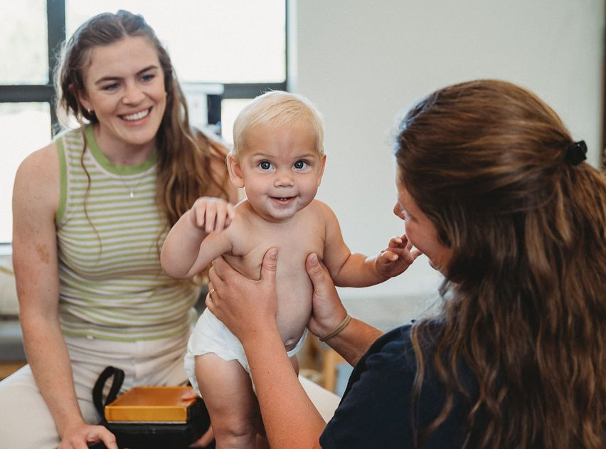 Woman holding smiling baby while another woman sits nearby in a bright room