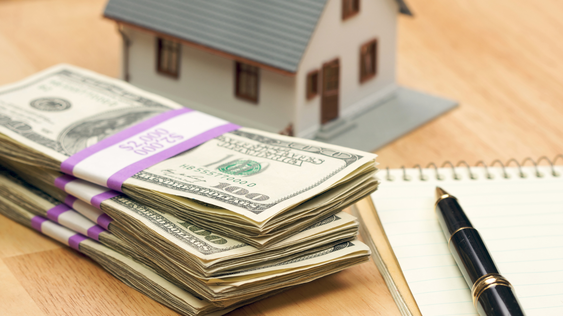A stack of money is on a table next to a pen and a model house.
