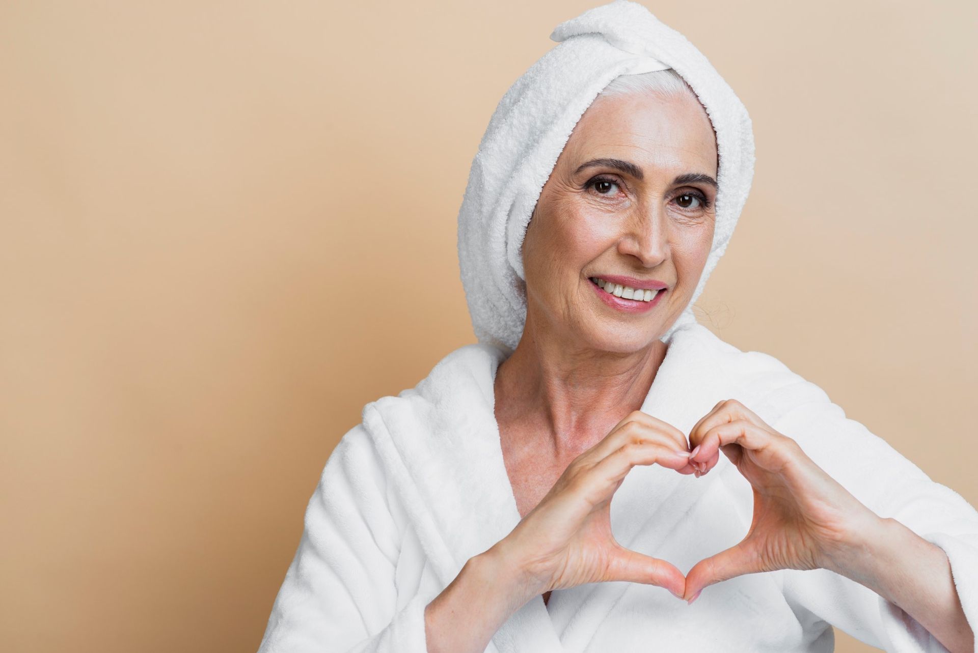 Woman in a white robe and towel on her head, smiling and making a heart shape with her hands.