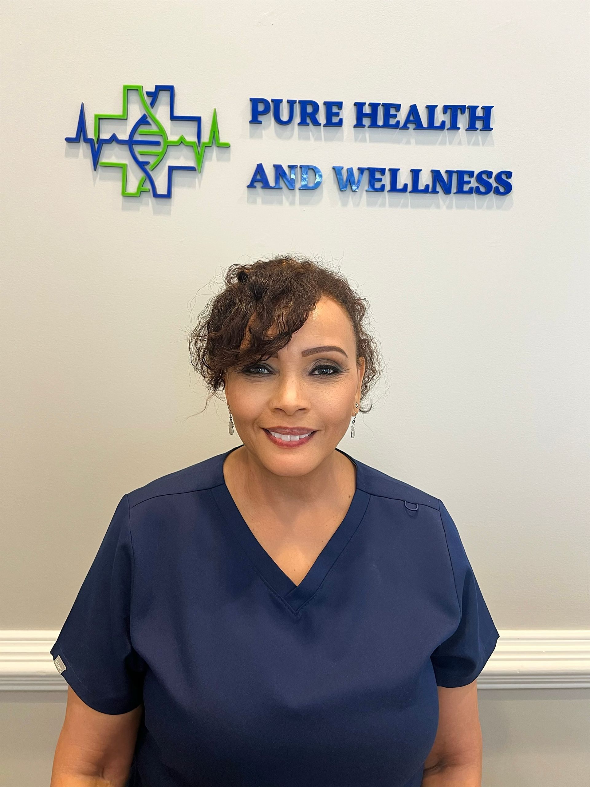 Woman in navy scrubs standing before a Pure Health and Wellness sign, smiling.