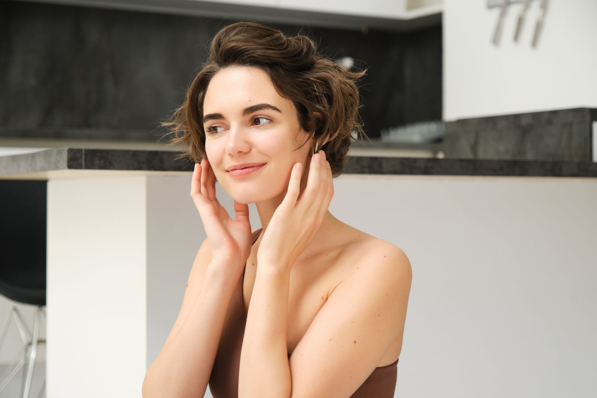 A smiling person with short brown hair touching their cheeks in a bright, modern kitchen.