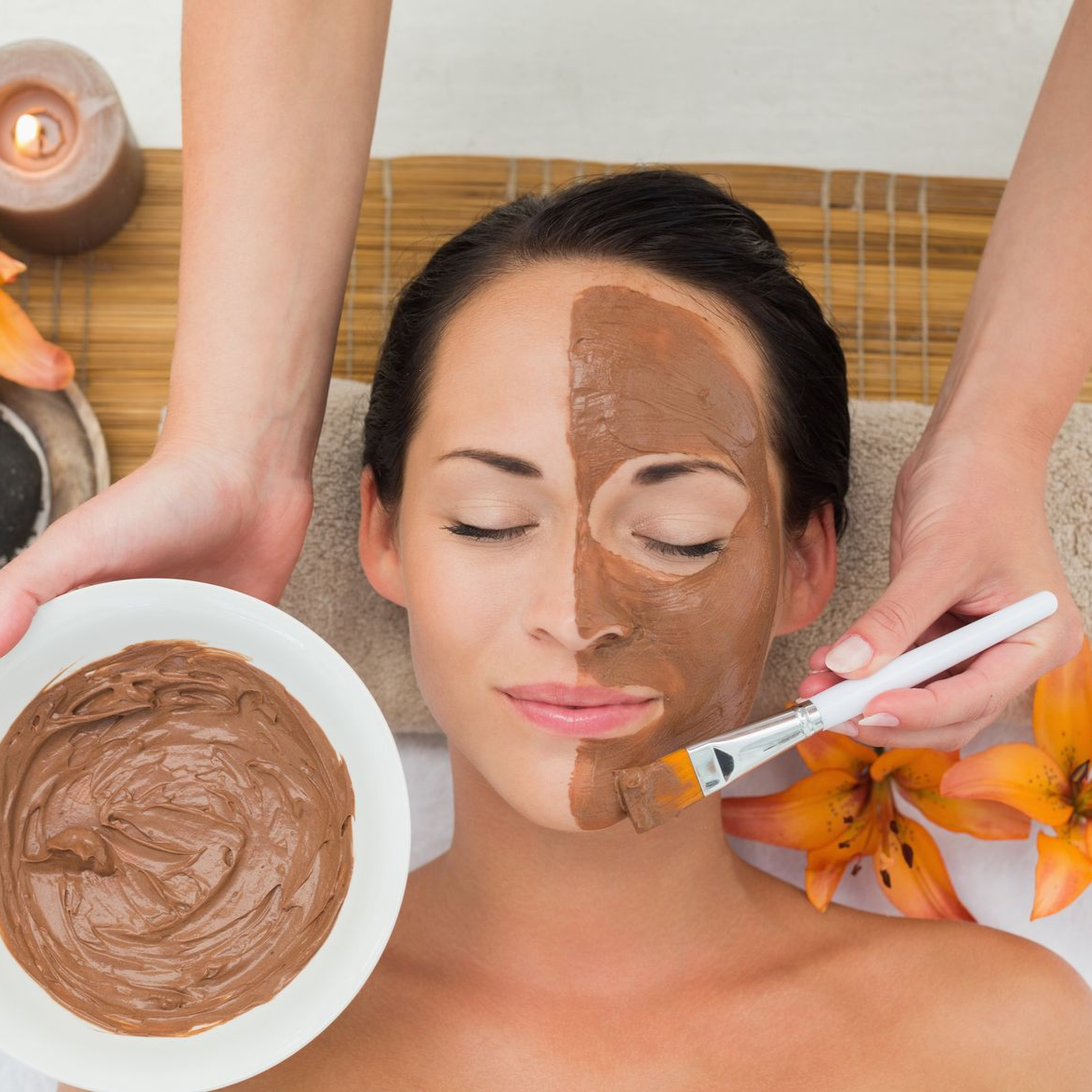 A spa professional applies a brown clay face mask with a brush to a person lying on a table beside candles and flowers.