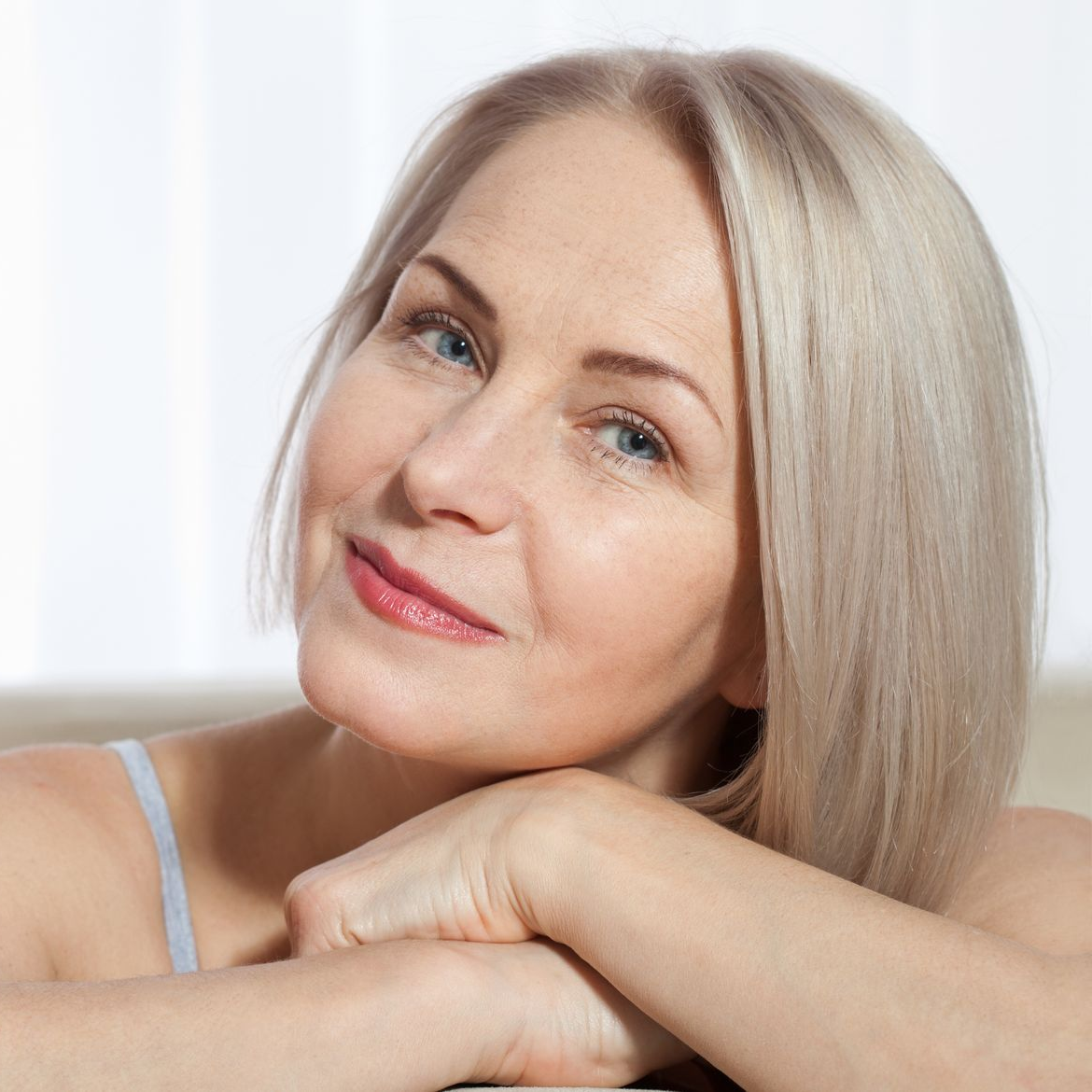 A person with light hair and blue eyes rests their chin on their hands, smiling softly against a bright, blurred backdrop.