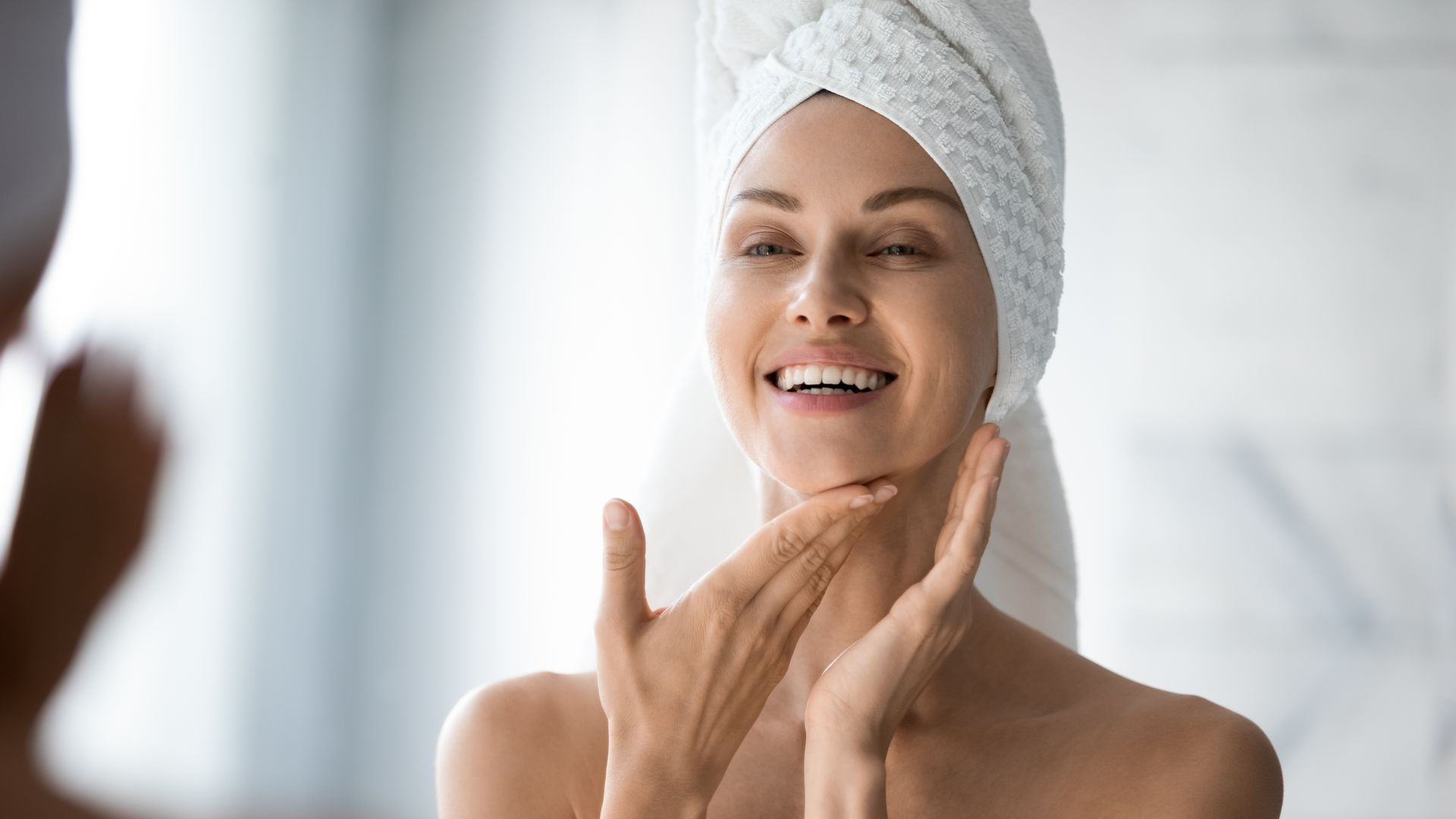 A smiling person with a towel wrapped around their head gently touches their chin while looking in a mirror.