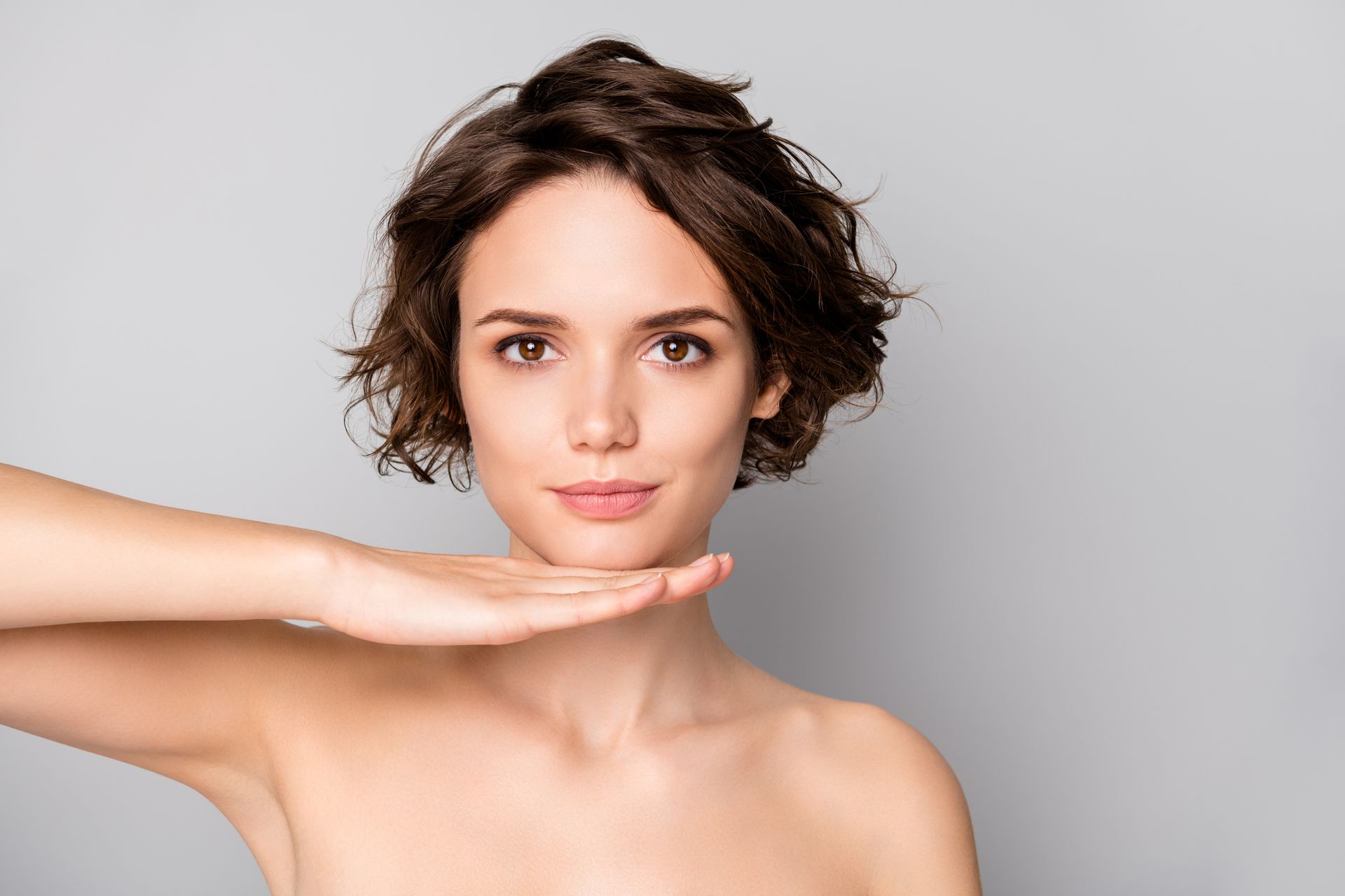 A person with short, wavy brown hair resting their chin on their hand against a plain gray background.