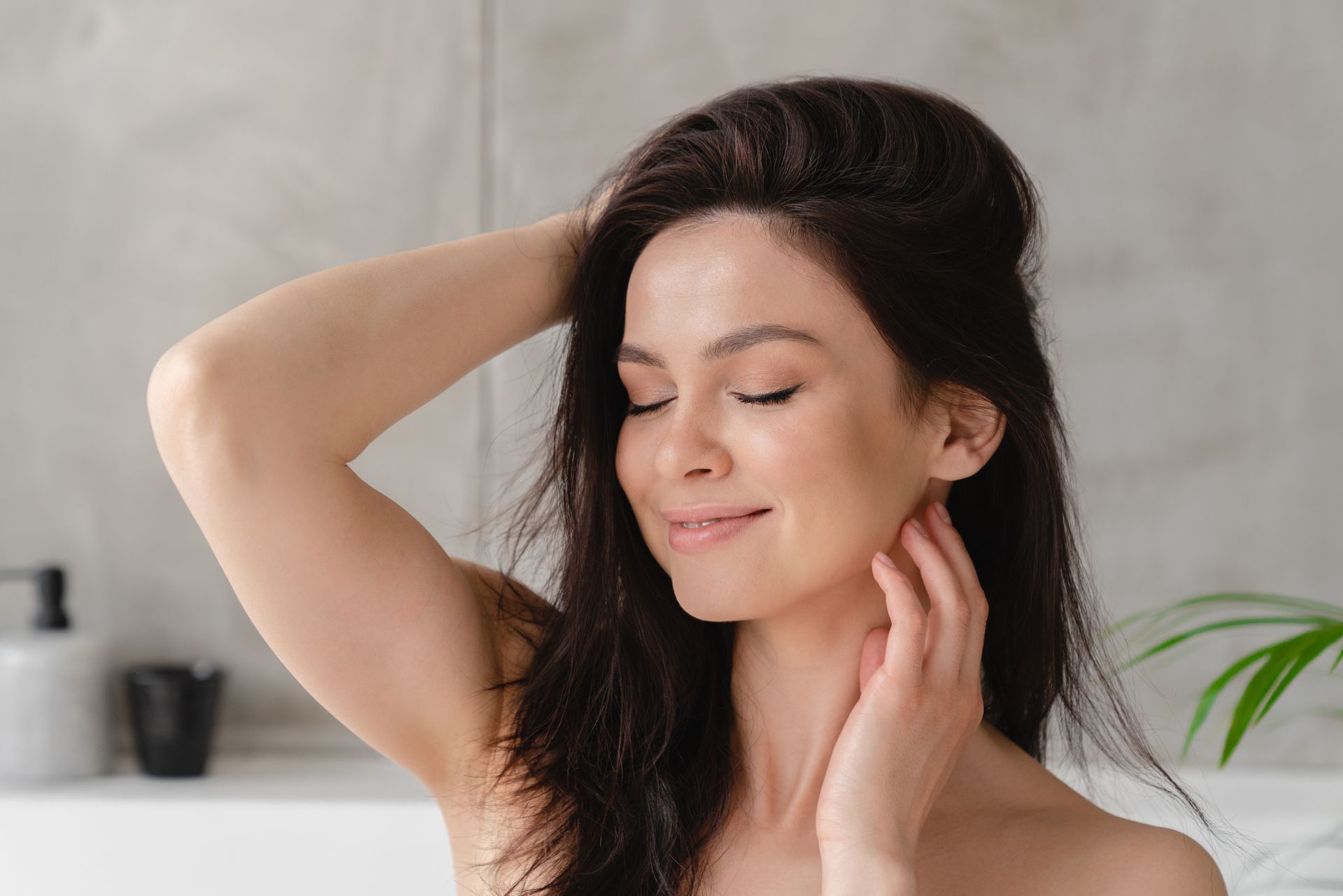 A person with dark hair closes their eyes and smiles, touching their neck while sitting in a light, neutral-toned space.