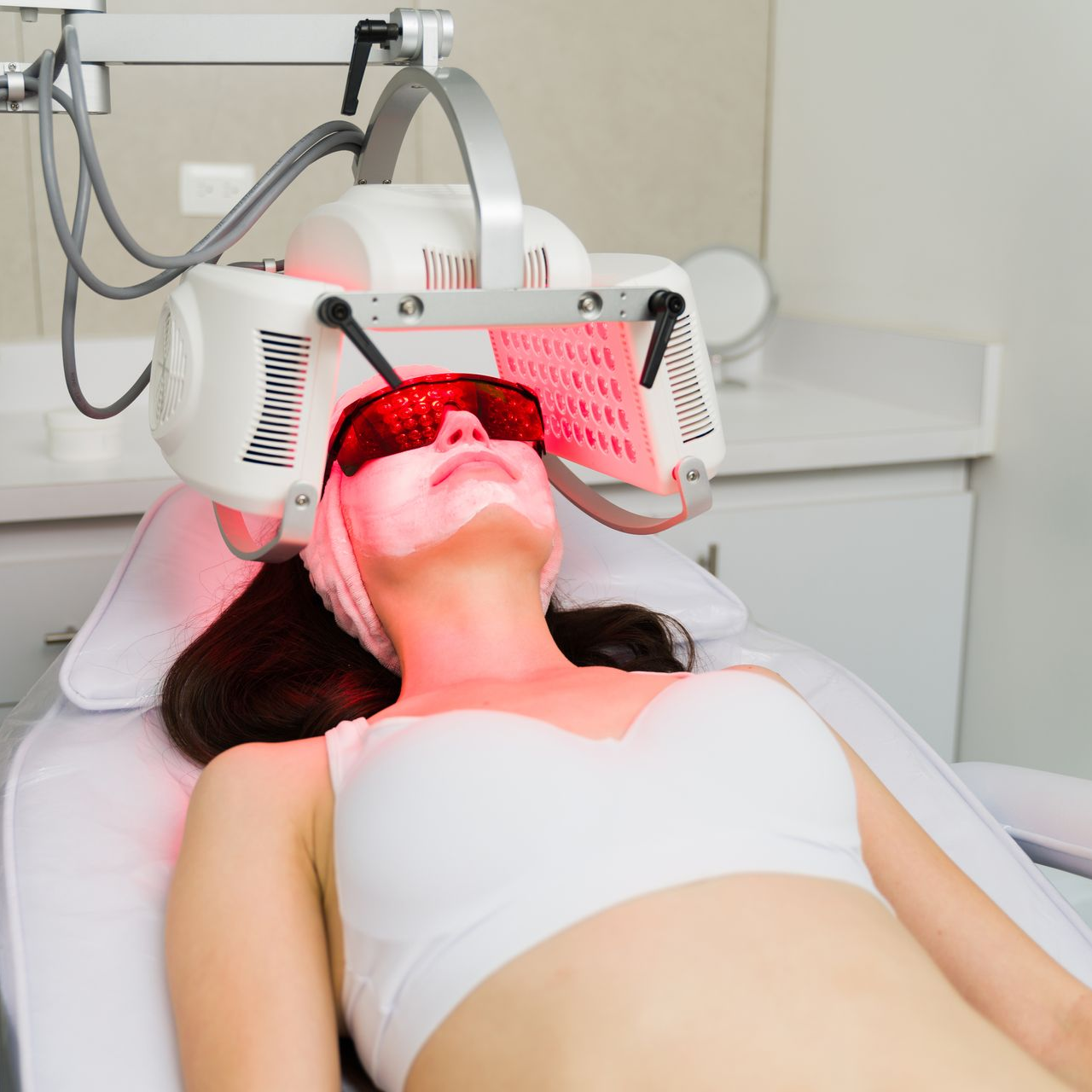 A person lying on a spa bed receives red light therapy treatment while wearing protective red goggles.