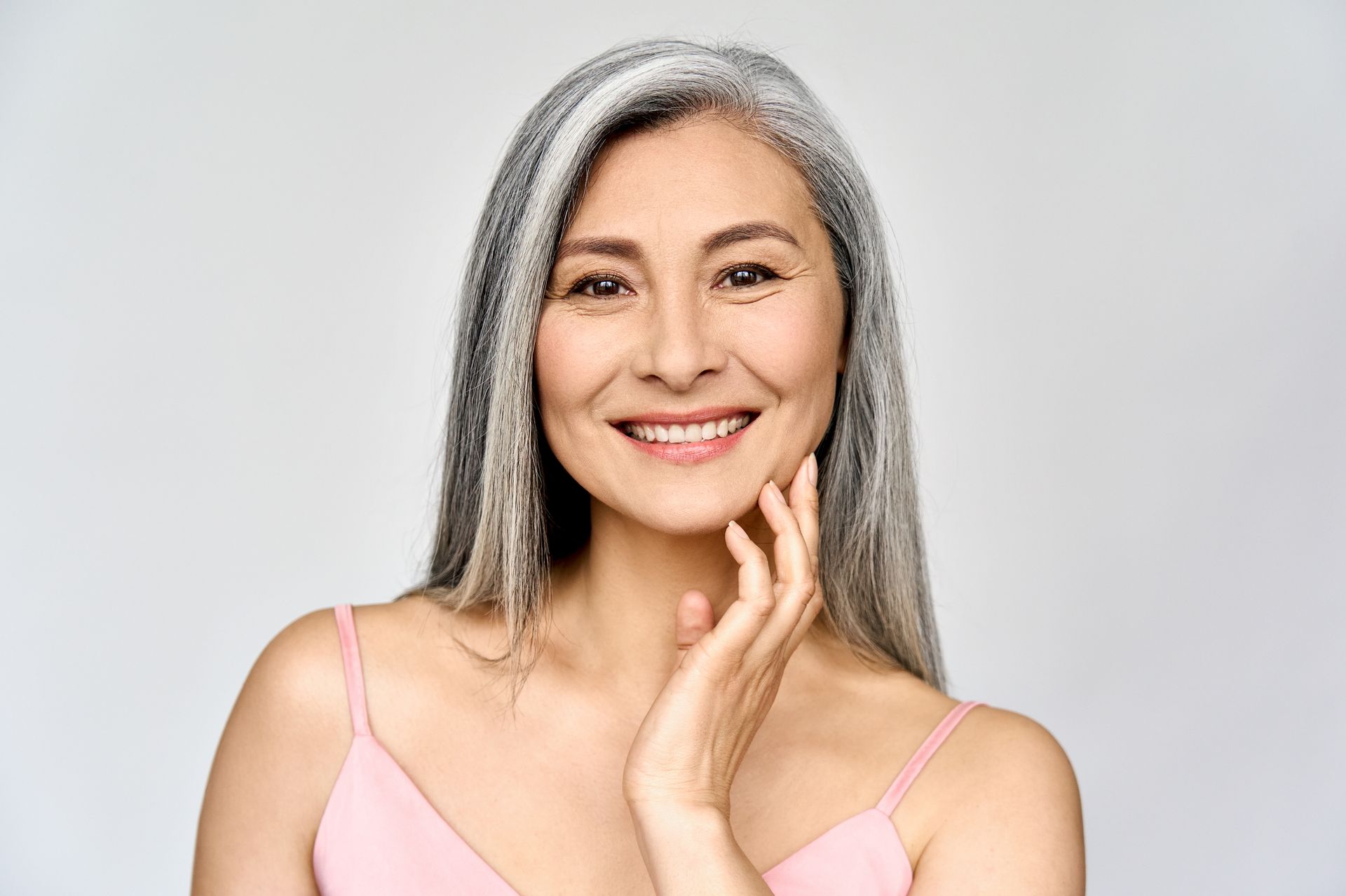 Smiling woman with long gray hair in a pink top, resting her hand on her chin against a light gray background