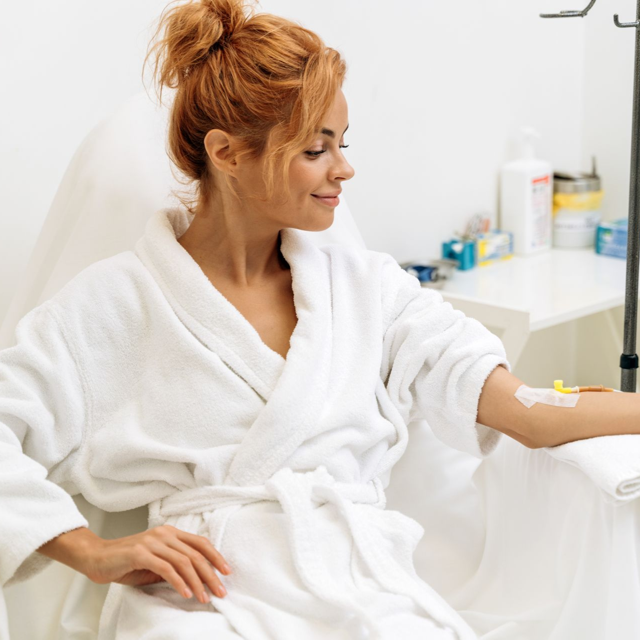 A person wearing a white robe sits in a clinic chair with an intravenous line in their arm, looking toward their hand.