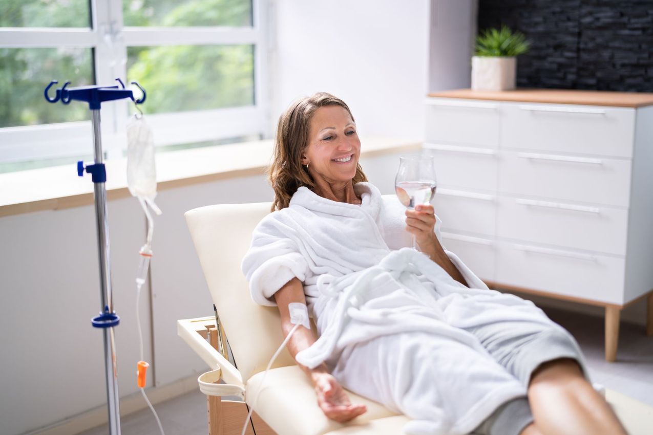 A person in a white robe receives an IV drip while relaxing in a spa setting, holding a glass of water.