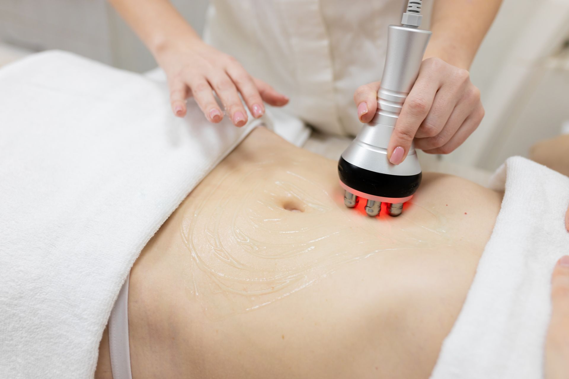 A technician uses a handheld radiofrequency device with red light therapy on a client's abdomen coated in conductive gel.