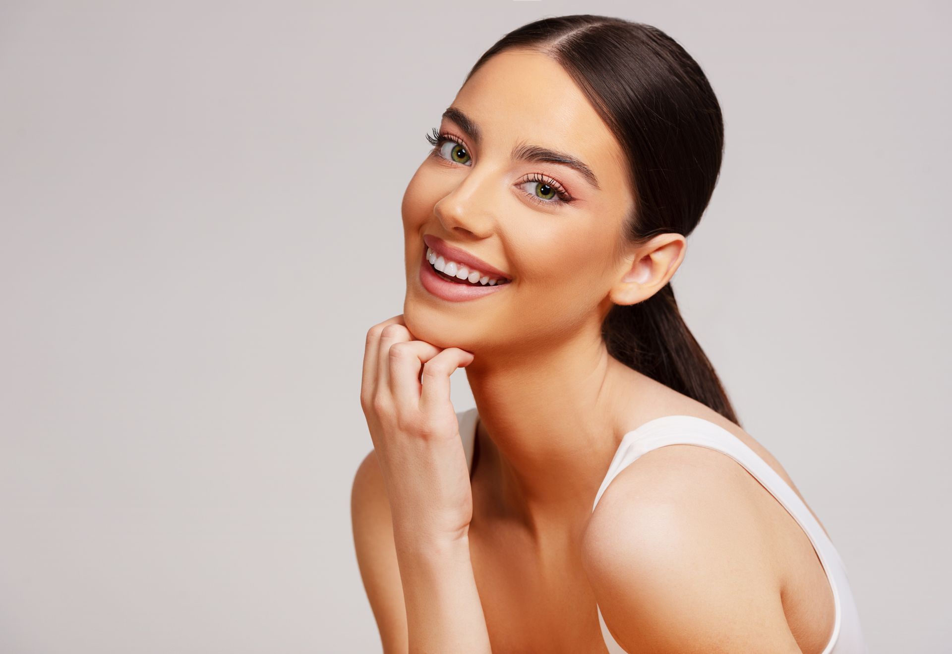 Smiling woman with dark hair in a white top, resting her chin on her hand against a light background