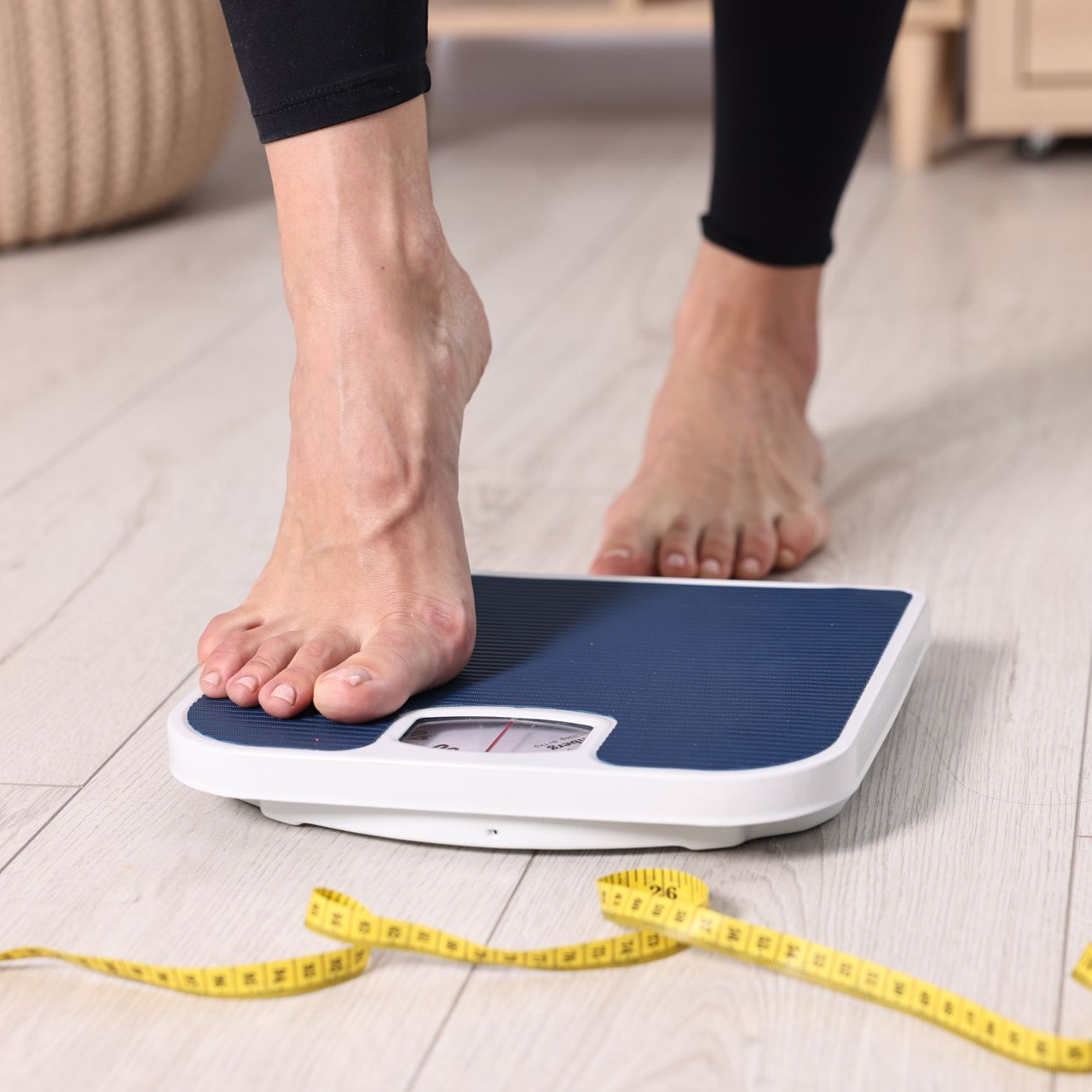 Feet on a bathroom scale beside a yellow measuring tape on a light floor