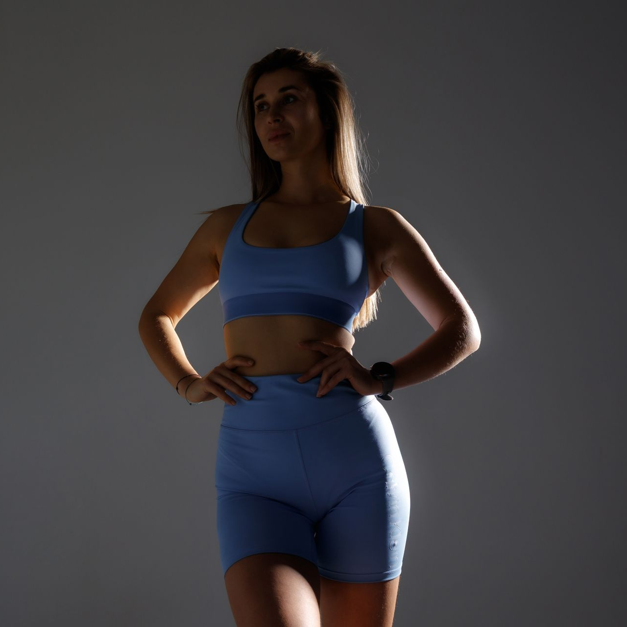A person with a fit physique wearing a black sports bra and shorts, posing with hands on hips against a gray background.