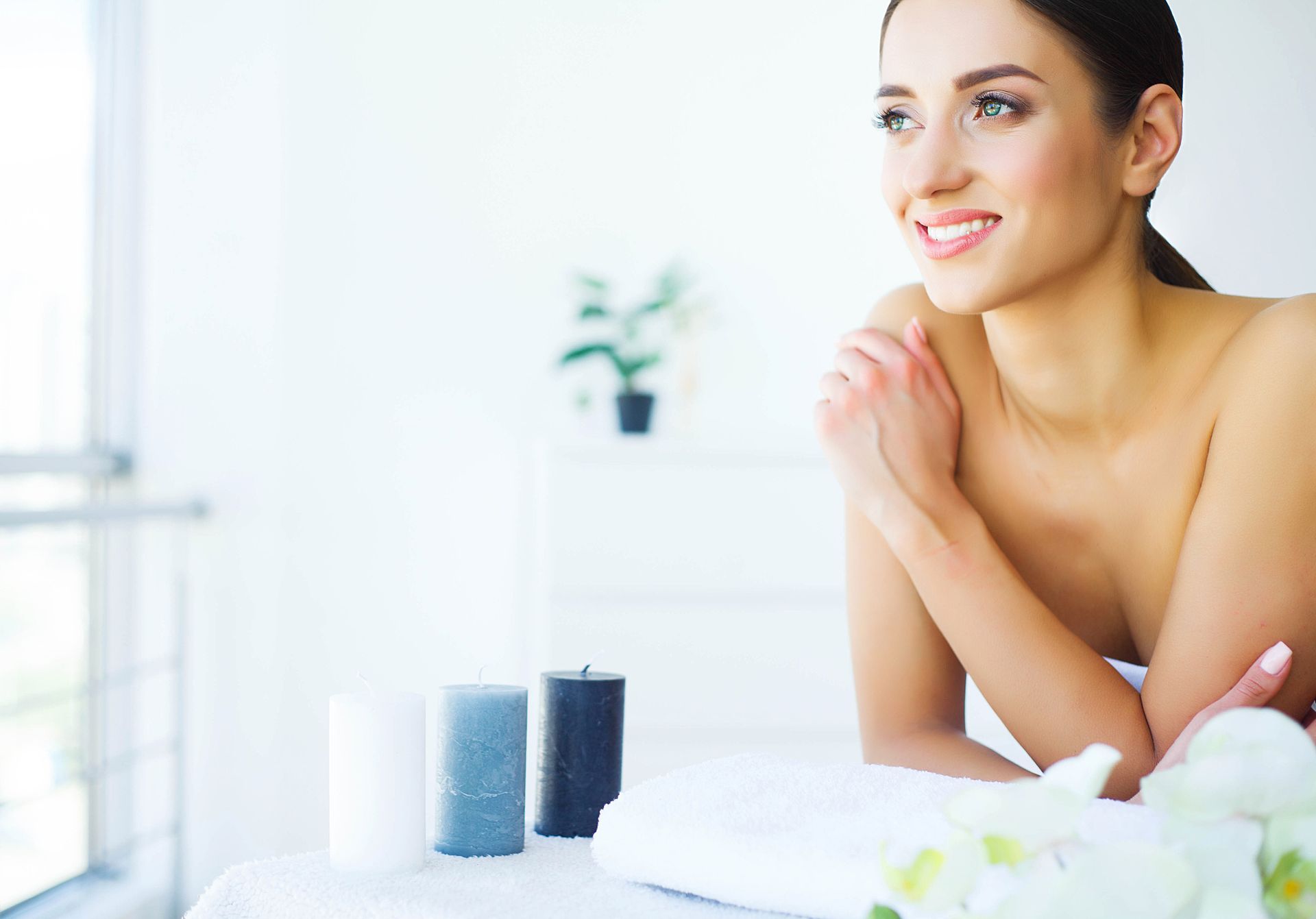 A person resting on a massage table with spa candles and flowers nearby in a bright, tranquil setting.