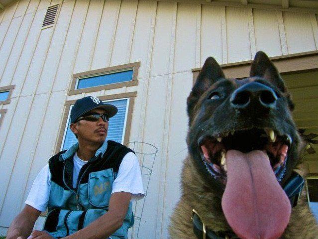 Man in cap and vest sits with a panting dog outside a light-colored house.