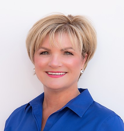 Blonde woman with a friendly smile, wearing a blue collared shirt, against a white backdrop.