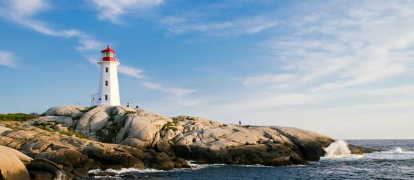 White lighthouse with a red top stands on a rocky outcrop, with blue sky and ocean in the background.
