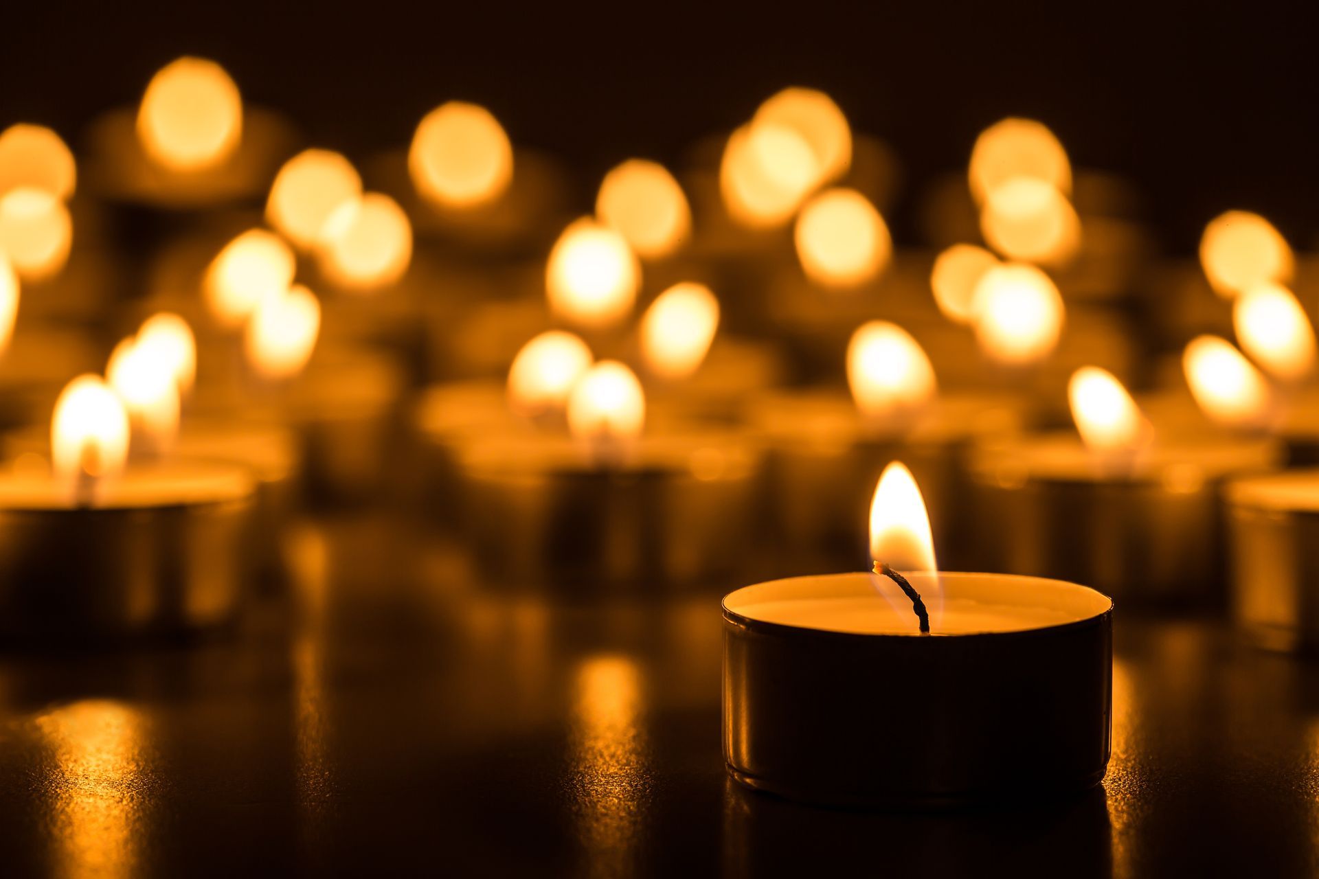 A single tea light candle glows in the foreground with many other blurred candles creating a warm, golden backdrop.