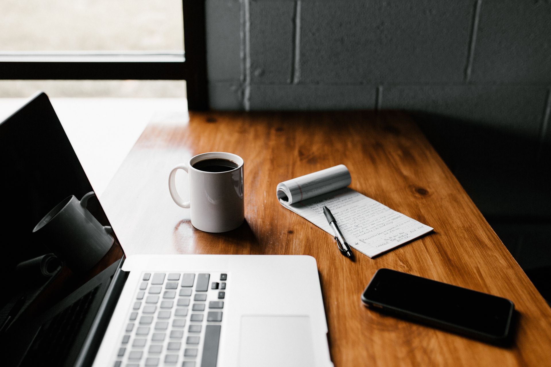 A laptop, coffee mug, notepad, pen, and smartphone sit on a wooden desk near a window.