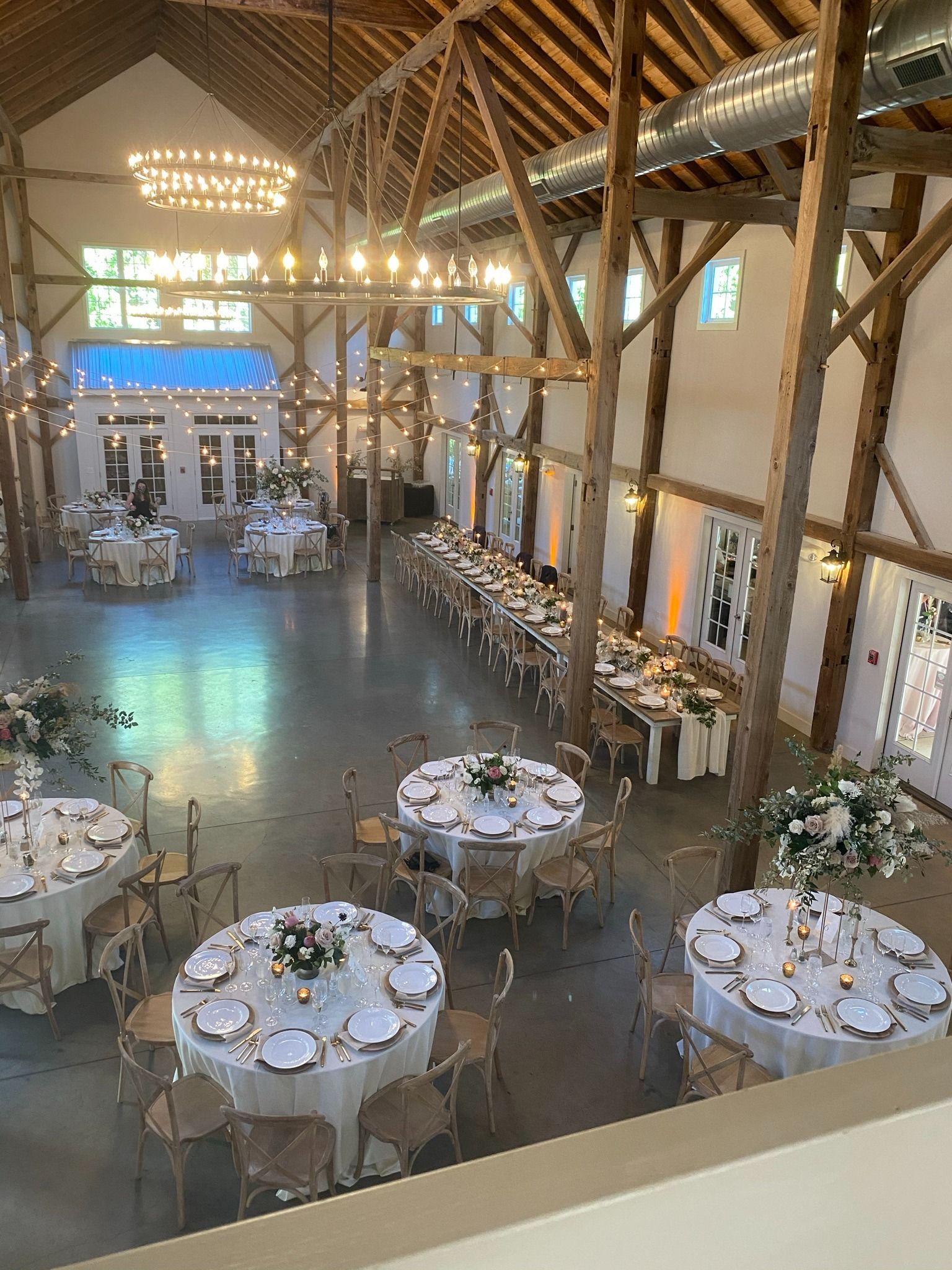 Rustic banquet hall with round tables, white chairs, tall wooden beams, and warm string lights