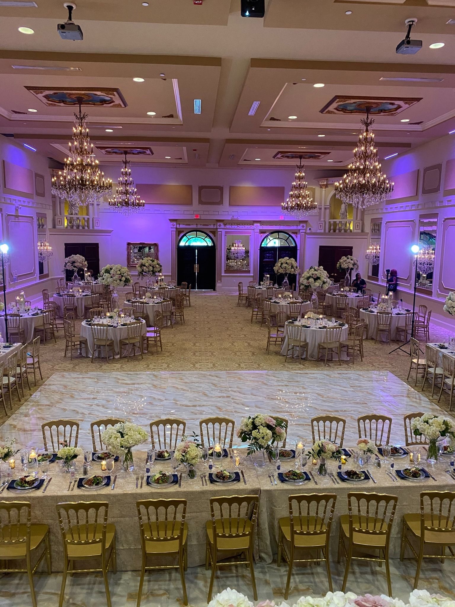 Elegant banquet hall with round tables, gold chairs, floral centerpieces, and purple accent lighting