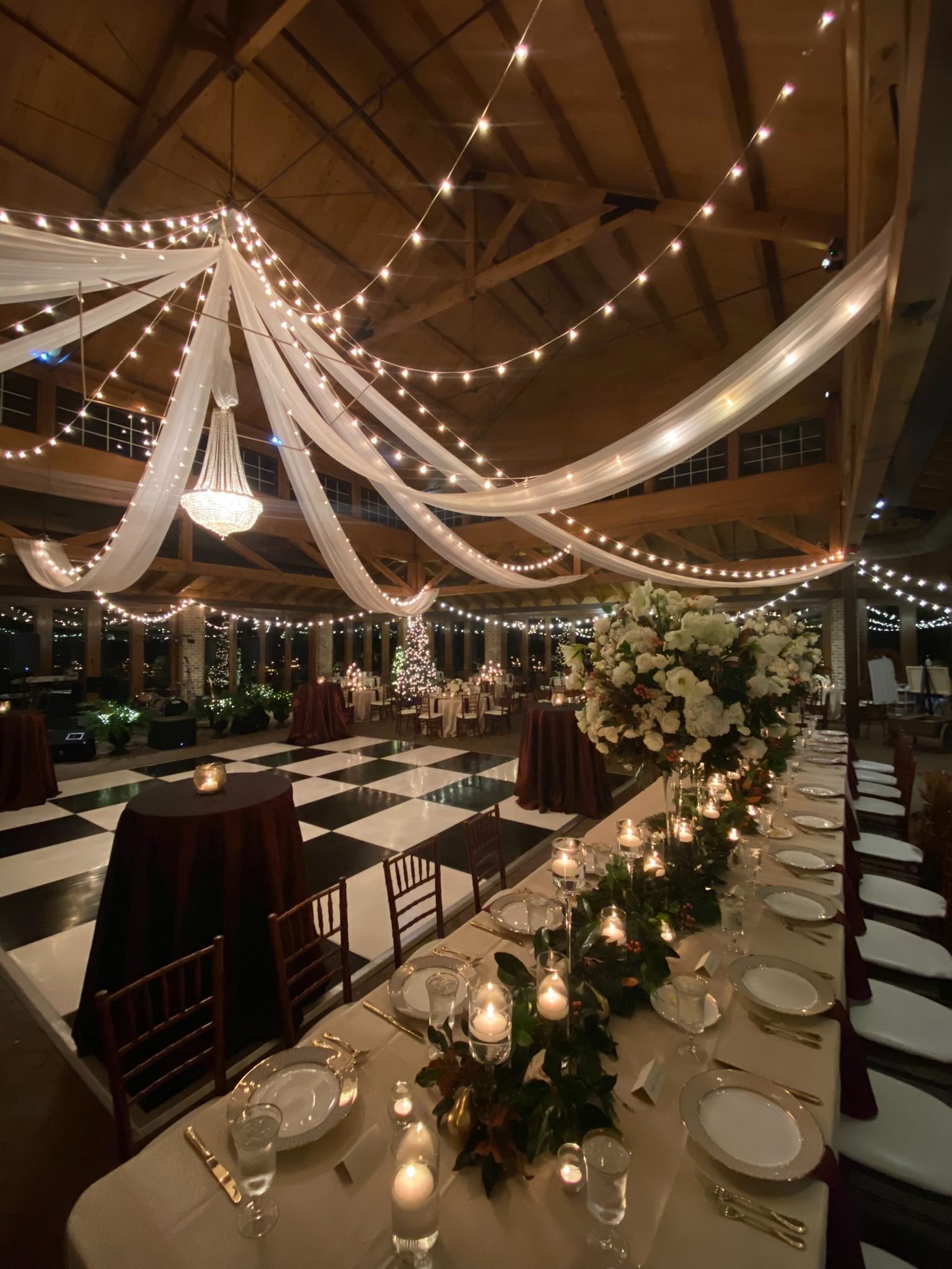 Elegant indoor banquet hall with draped white fabric, string lights, and candlelit tables decorated with flowers.