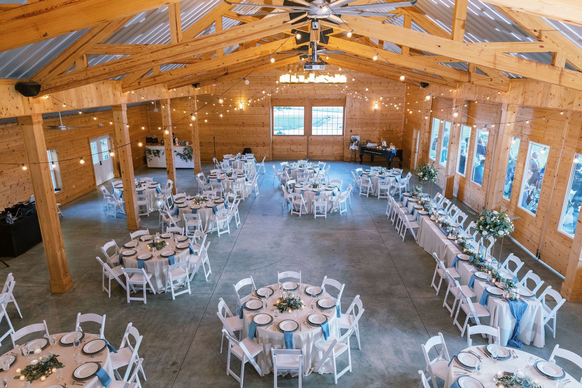 Wooden banquet hall with round tables, white chairs, and string lights, set for an event