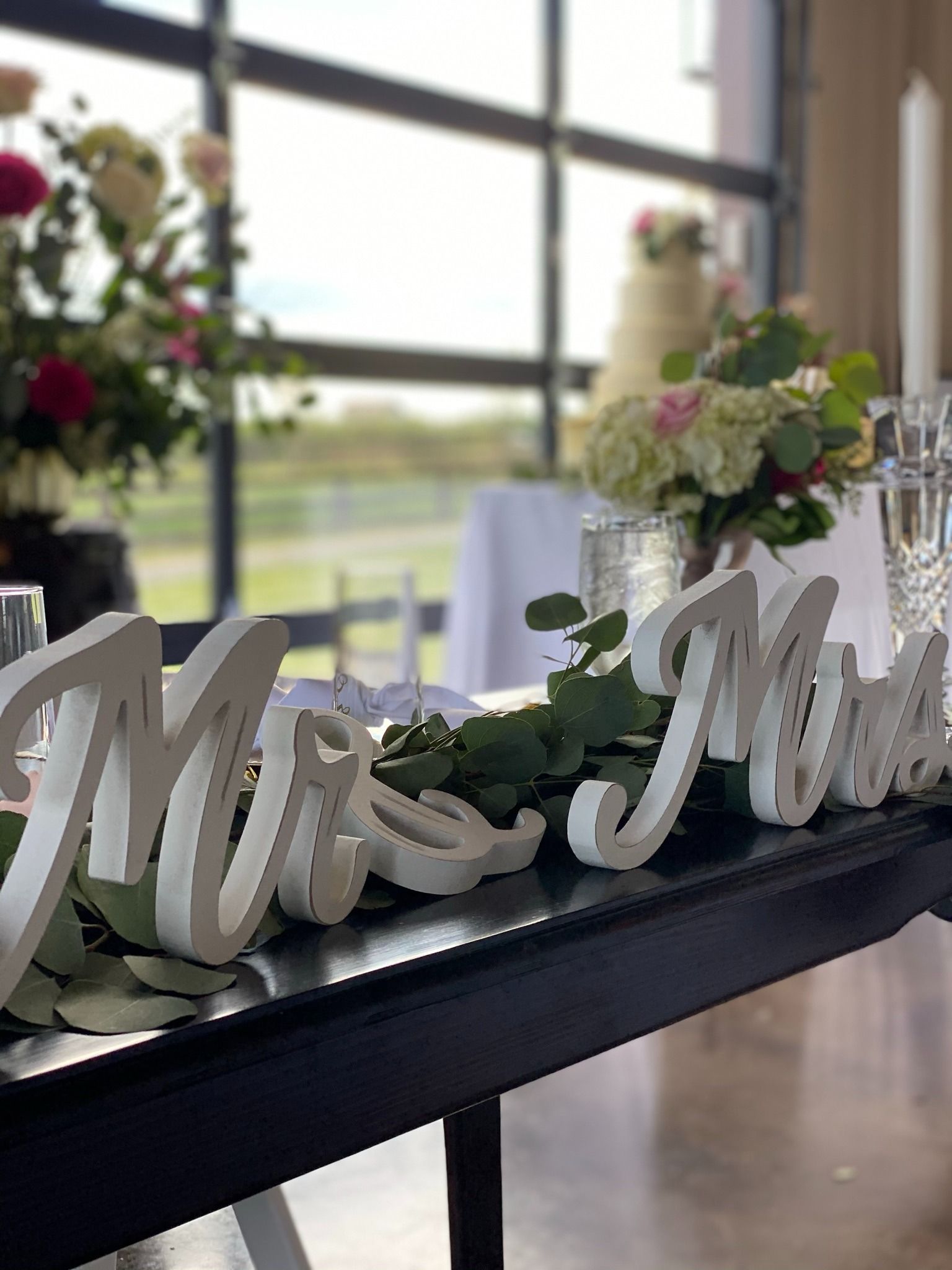 Decorative “Mr & Mrs” sign on a table with flowers at a wedding reception near large windows