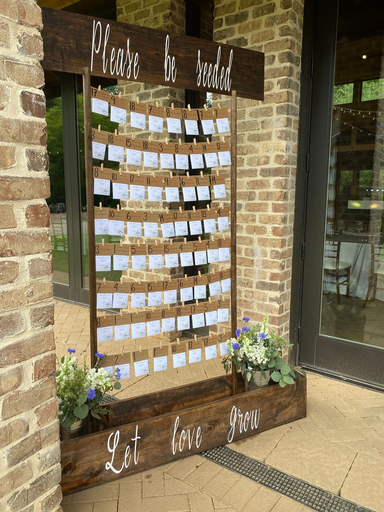 Wooden prayer/tribute display with many notes, set against a brick wall and glass doors, with flowers at the base