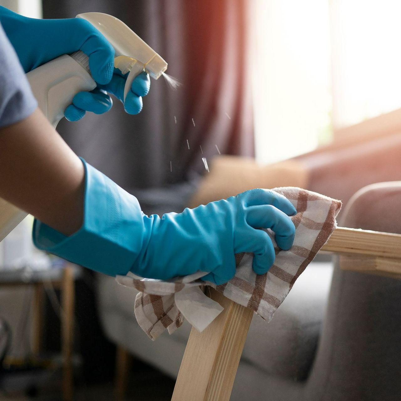 A person wearing blue gloves is cleaning a wooden table with a spray bottle.