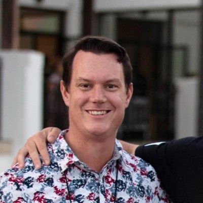 Man in patterned shirt smiling, arm around someone, outdoors.