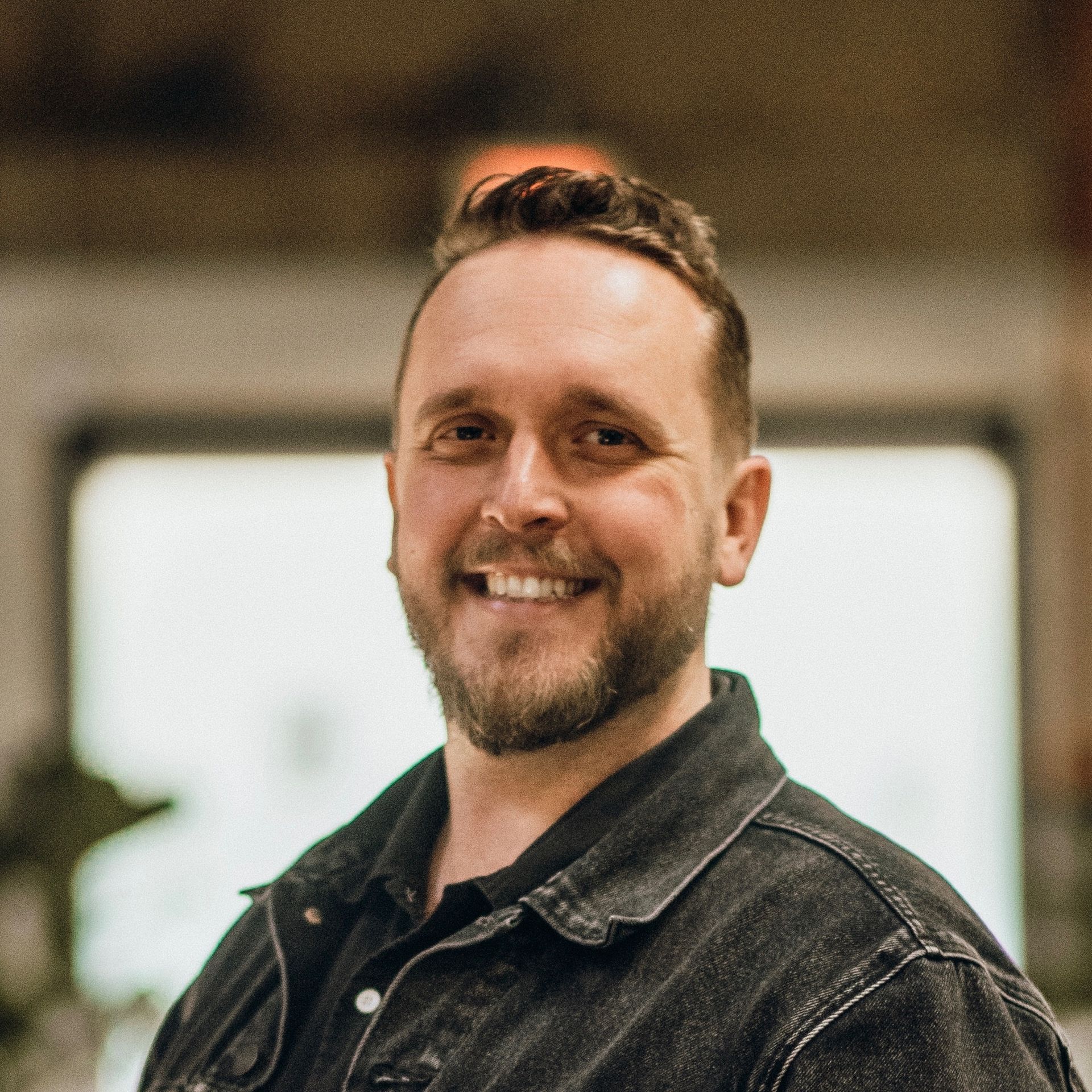 Man with a beard smiling, wearing a black denim jacket, indoors.