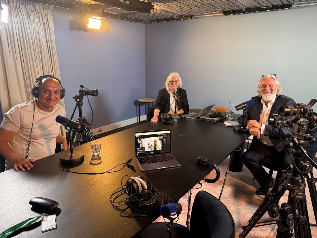 Drie mensen maken een podcast in een blauwe opnamestudio met microfoons en een laptop op tafel.