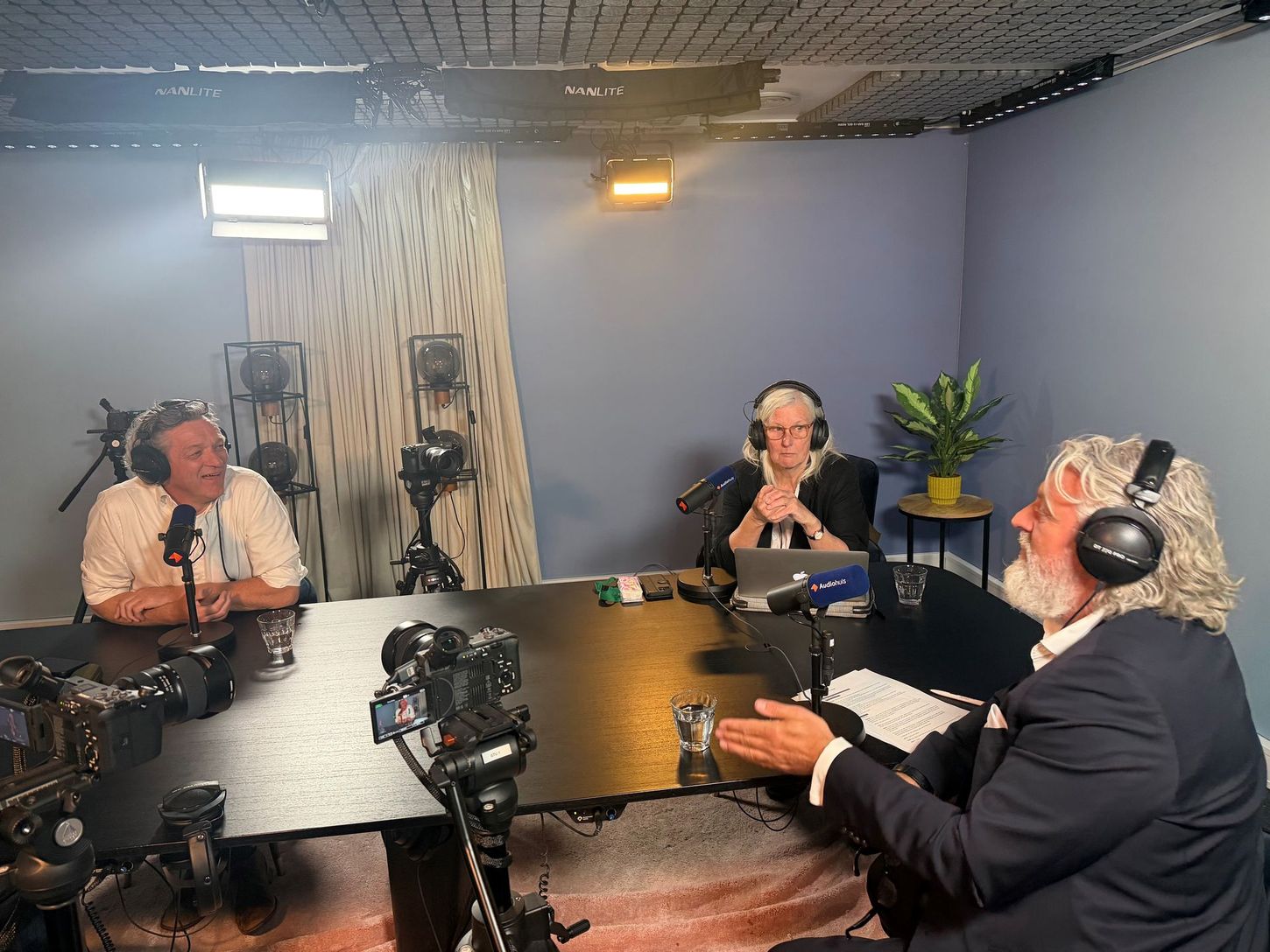 Drie mensen maken een podcast in een blauwe opnamestudio met microfoons en een laptop op tafel.