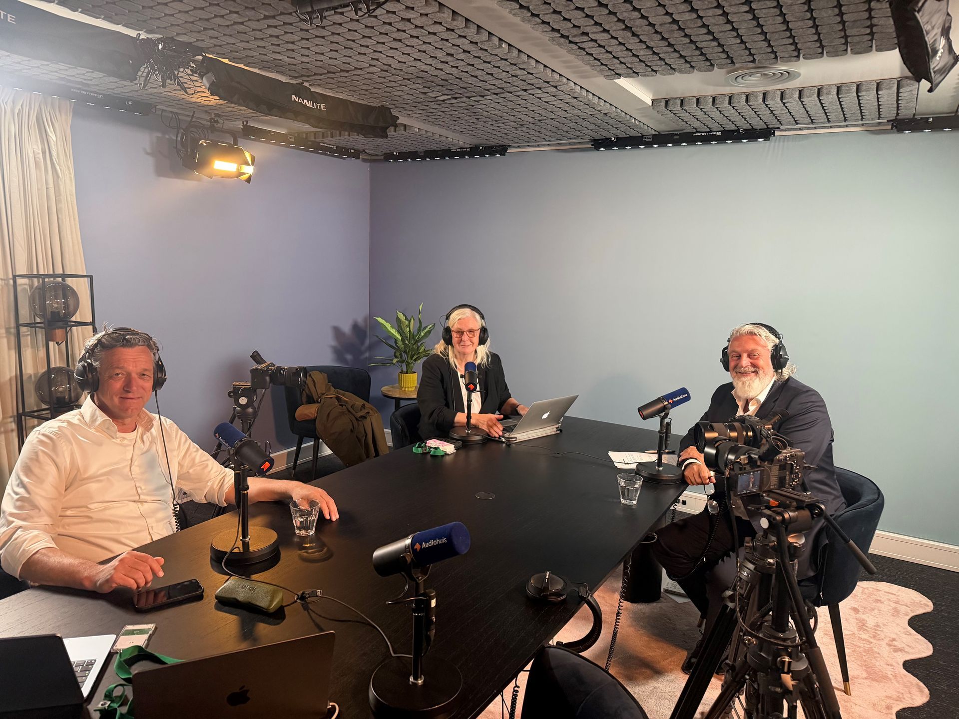 Drie mensen maken een podcast in een blauwe opnamestudio met microfoons en een laptop op tafel.