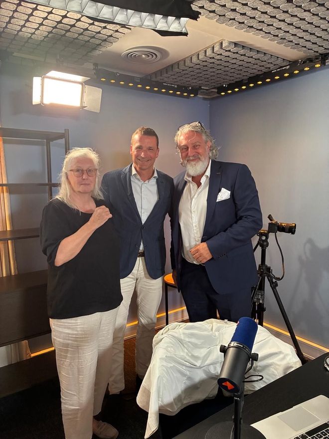Drie mensen maken een podcast in een blauwe opnamestudio met microfoons en een laptop op tafel.