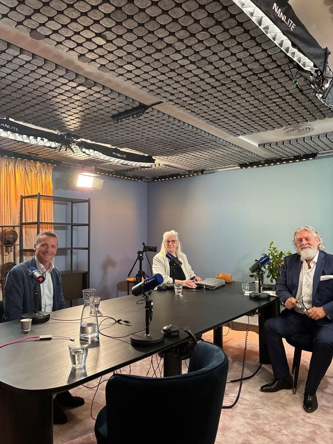 Drie mensen maken een podcast in een blauwe opnamestudio met microfoons en een laptop op tafel.