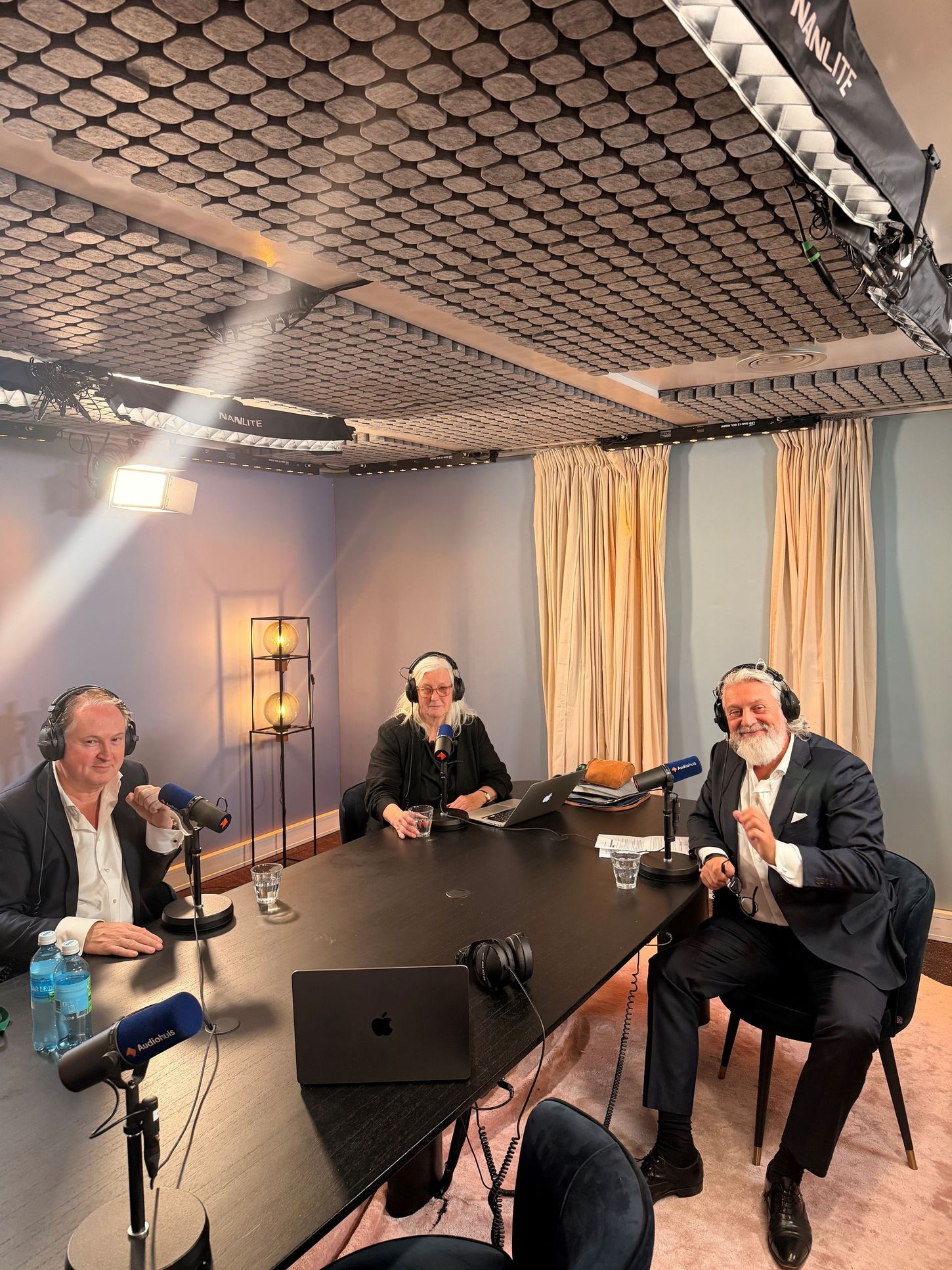 Drie mensen maken een podcast in een blauwe opnamestudio met microfoons en een laptop op tafel.