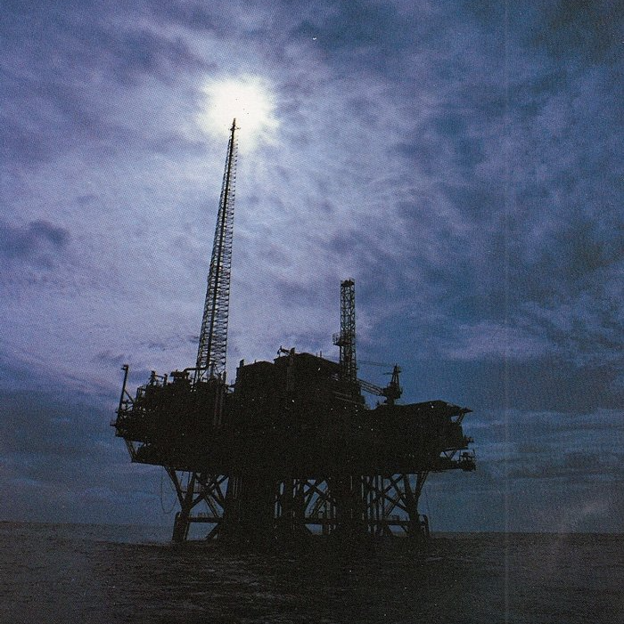 A silhouette of an oil rig with the sun shining through the clouds
