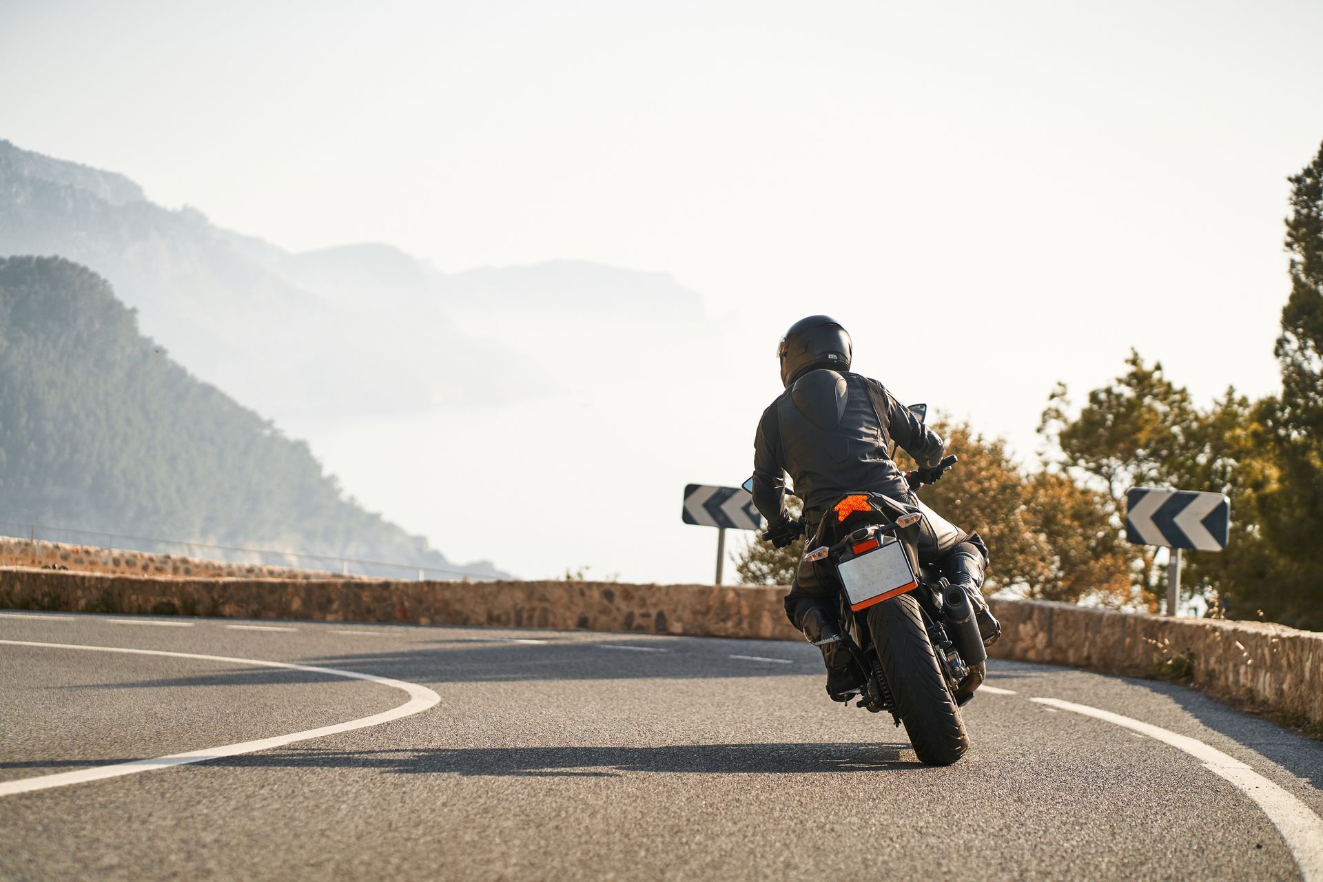 Motorcyclist leans into a turn on a winding road, mountains in the background, sunny day.