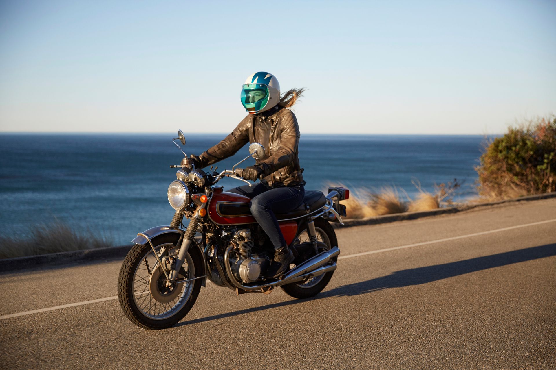 Woman in a helmet rides a vintage motorcycle on a coastal road with the ocean in the background.