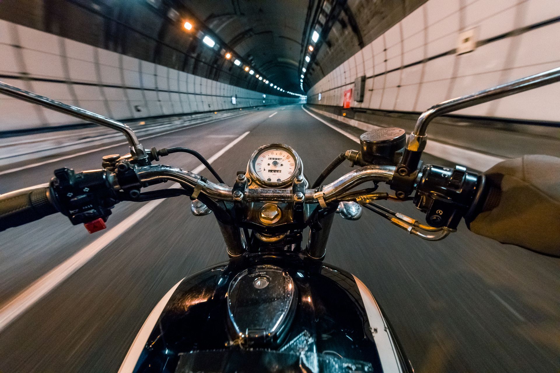 Motorcycle speeding through a long tunnel. The rider's view shows the handlebars and dashboard, with lights illuminating the road ahead.