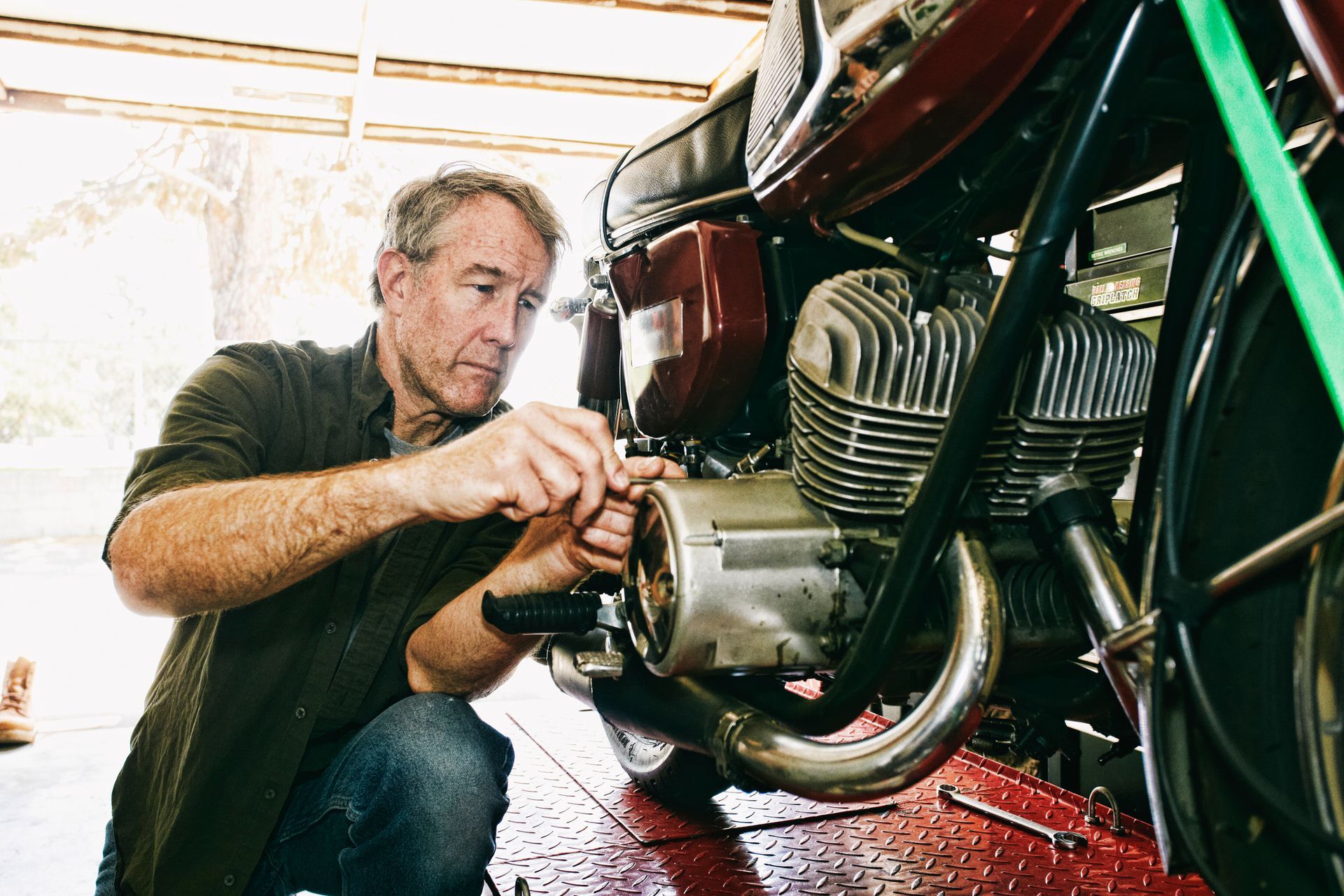 A man working on a motorcycle engine in a garage. He kneels, focused, using a tool.